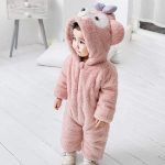 গোলাপি bear onesie for toddlers পরা একটি শিশু ঘরের ভিতরে দাঁড়িয়ে আছে — ফাজি ও হুডেড ডিজাইন সহ।